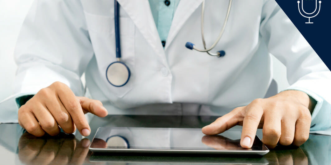 clinician in white coat with stethoscope around neck sitting at table and working on tablet