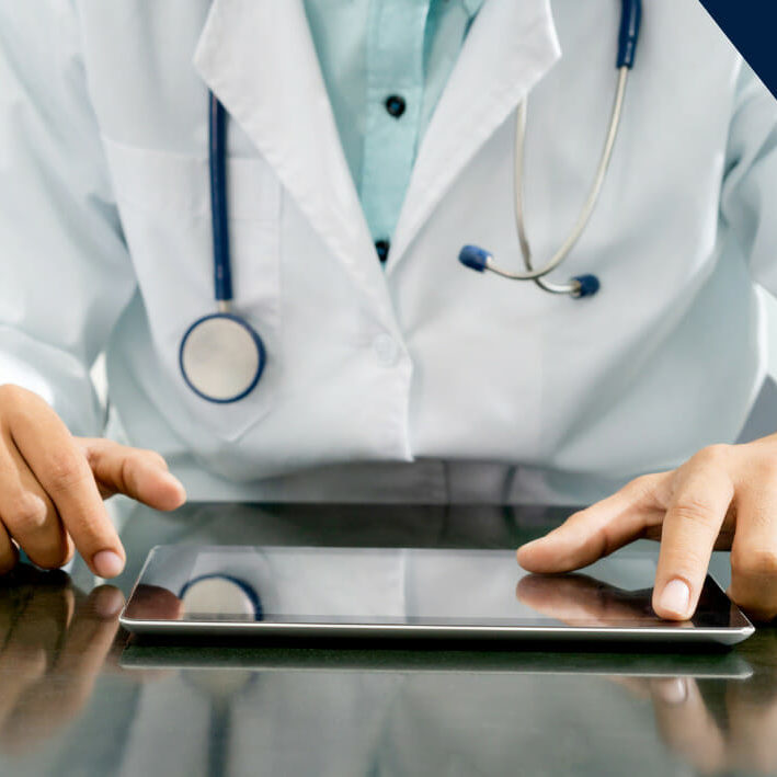 clinician in white coat with stethoscope around neck sitting at table and working on tablet