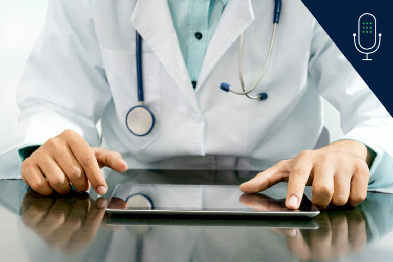 clinician in white coat with stethoscope around neck sitting at table and working on tablet