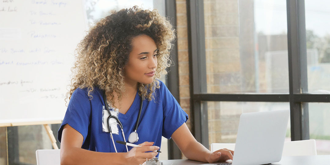 A nurse studying on a laptop while taking handwritten notes