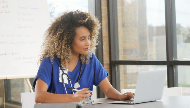 A nurse studying on a laptop while taking handwritten notes