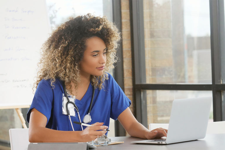 A nurse studying on a laptop while taking handwritten notes