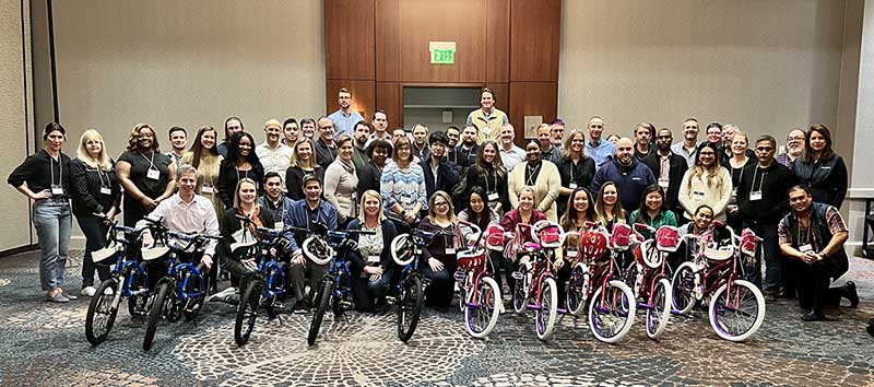 Approximately 50 uPerform employees pose with 10 children's bicycles in a large conference room.