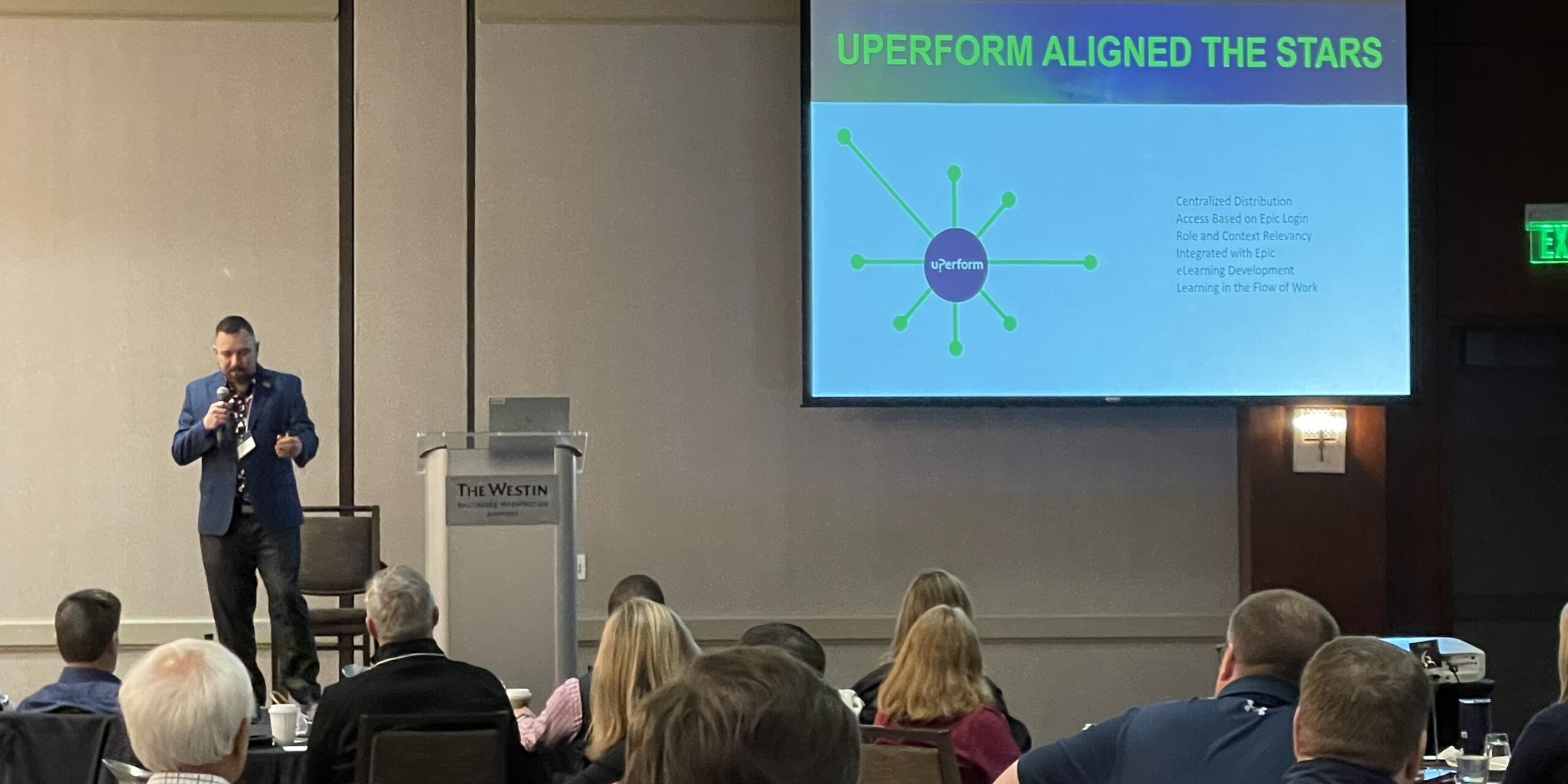 Derek Harley stands stage left speaking to audience with slideshow in top right of photo frame that reads "UPERFORM ALIGNED THE STARS" with some graphics and small supporting text.