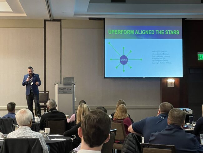 Derek Harley stands stage left speaking to audience with slideshow in top right of photo frame that reads "UPERFORM ALIGNED THE STARS" with some graphics and small supporting text.