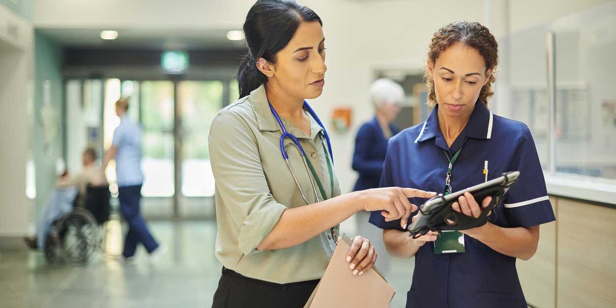 Female physician wearing a stethoscope assists female nurse using tablet in hospital setting.