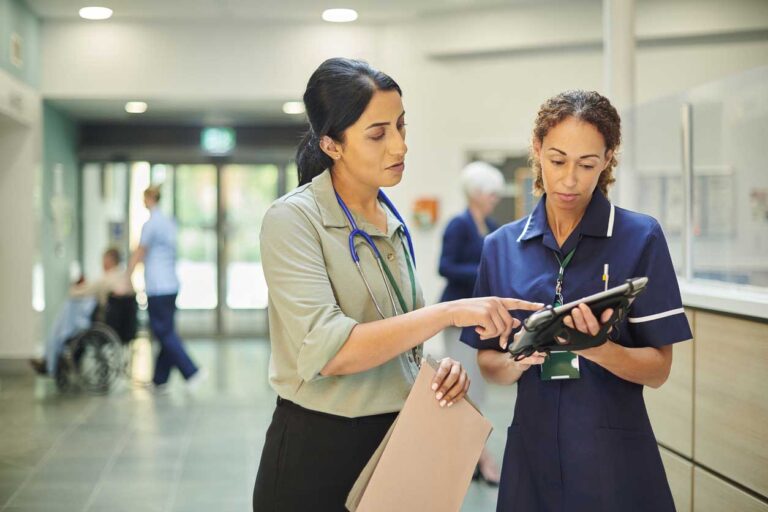 Female physician wearing a stethoscope assists female nurse using tablet in hospital setting.