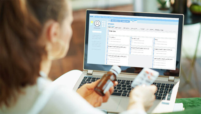 young female doctor is holding medication in front of an EHR screen while sitting down at laptop.