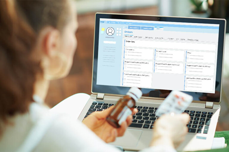 young female doctor is holding medication in front of an EHR screen while sitting down at laptop.