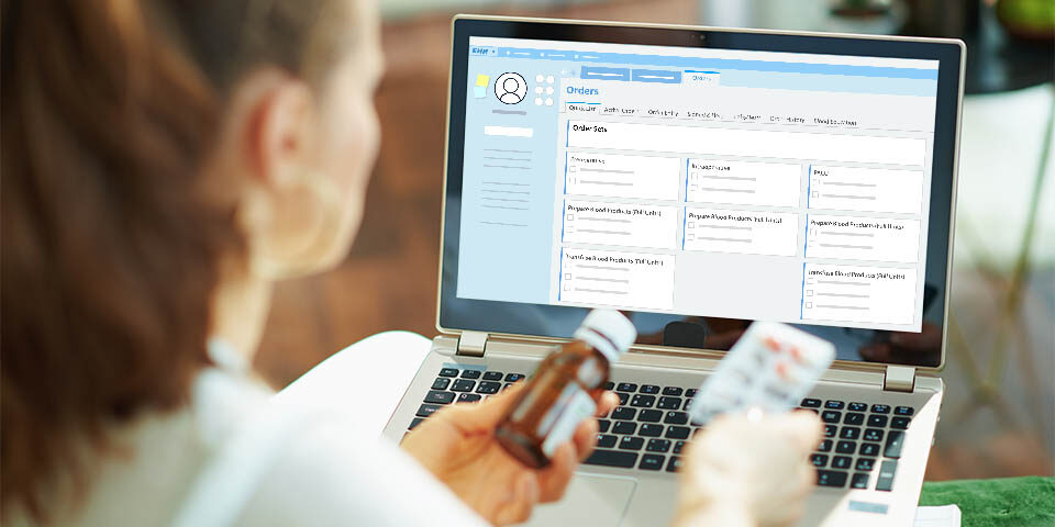 young female doctor is holding medication in front of an EHR screen while sitting down at laptop.