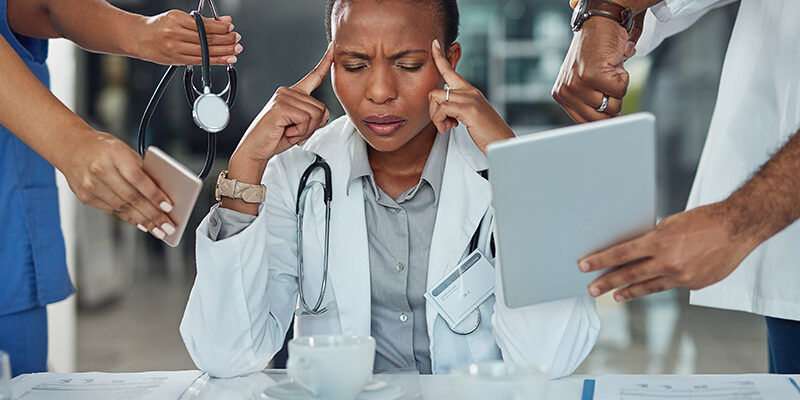 A stressed doctor presses her fingers to her temples, due to health worker burnout.