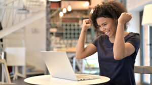 Woman sitting at table cheering with hands in the air looking at laptop