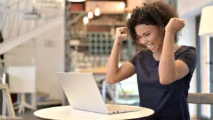Young African Woman Celebrating Success on Laptop in Cafe Woman sitting at table cheering with hands in the air looking at laptop