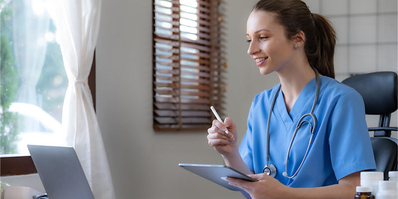 Female doctor focuses on laptop while she holds a tablet and stylus.