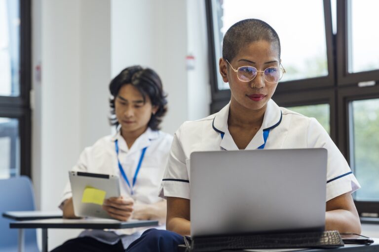 digital-adoption-platform-nhs-nurse-epr-learning Nurse using a laptop during a digital learning session, with a colleague reviewing a tablet in the background.